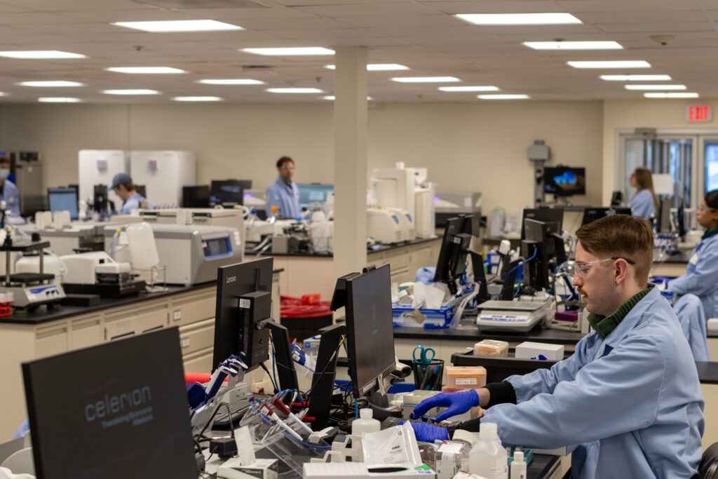 A photo of a lab. Tables with computer screens and lab equipment with people in blue lab coats and safety goggles. 